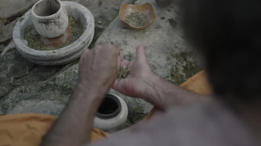 A man sits on the ground and puts a leafy substance in his palm