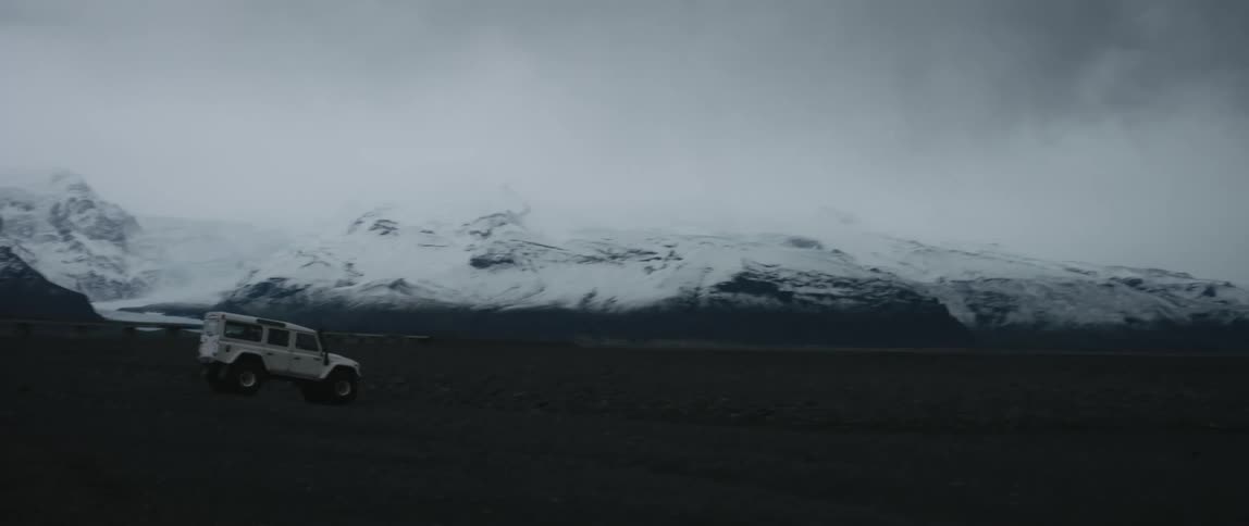 A person drives a jeep through a dirt field below snow covered mountains
