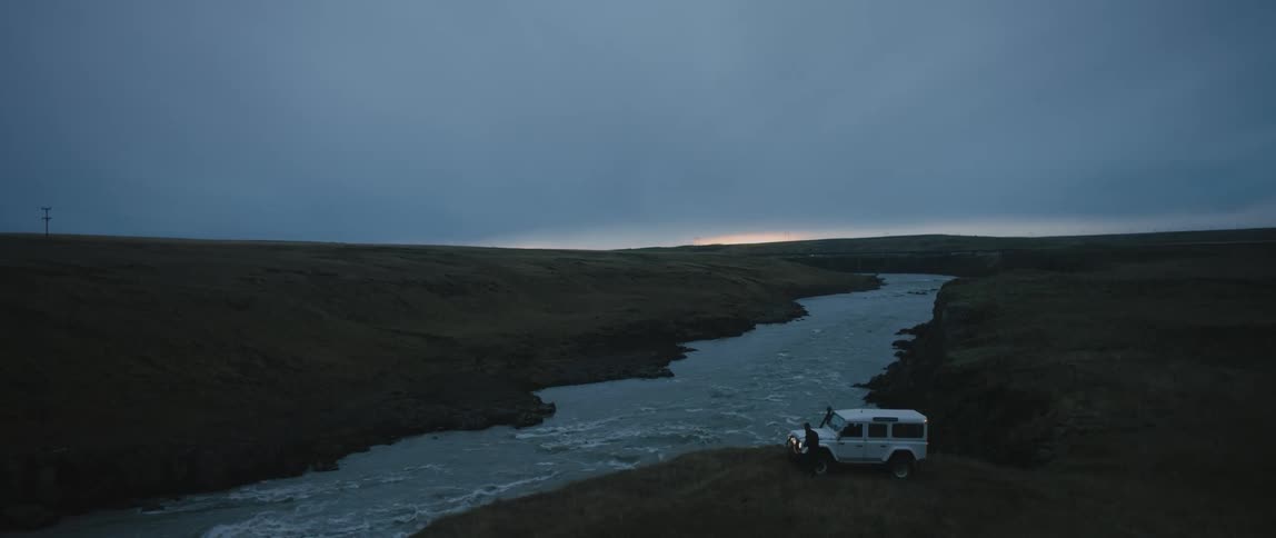 A person exits an offroad vehicle parked on the bank of a river on a cloudy evening
