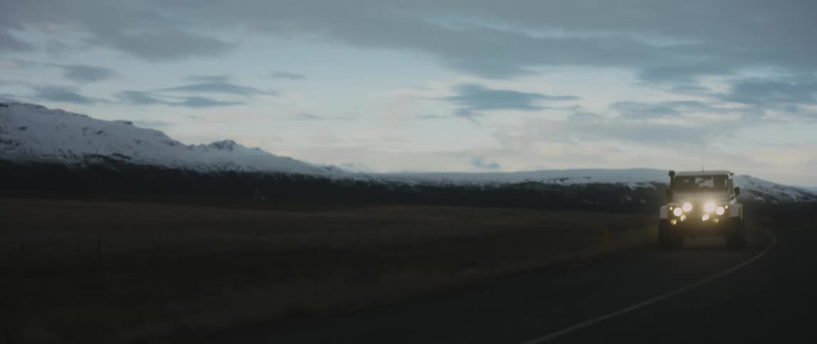 White vehicle travels on a highway at night next to a field with snowy mountains in the distance