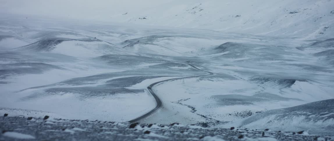 White vehicle travels on the highway between two snowy fields and with snowy mountains behind it