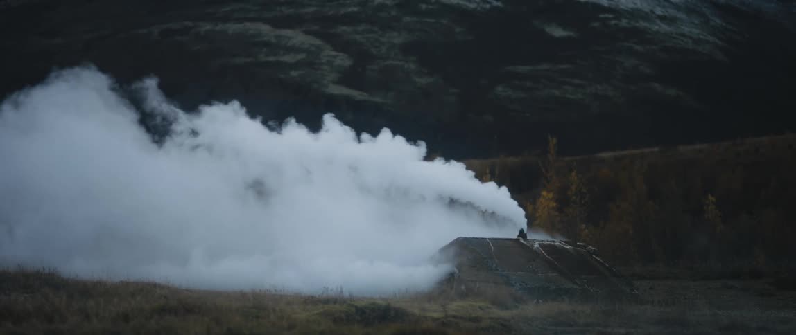 Smoke pours from small building in field near woods towered over by snow covered mountain