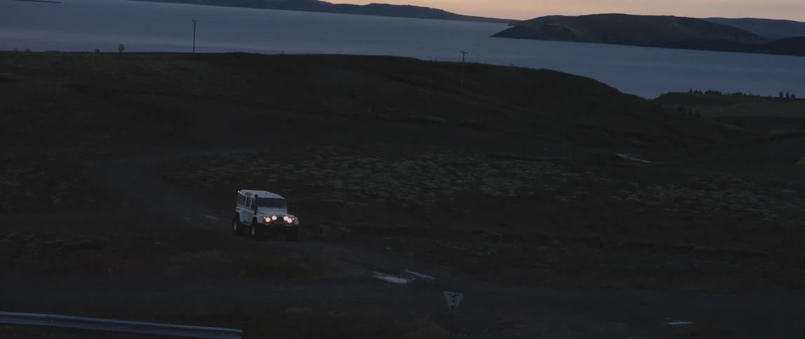 White vehicle travels on a dirt road near the hills and goes towards a paved highway