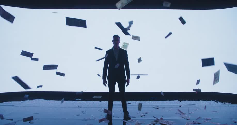 Silhouette of a young business man standing in front of a large white digital display while dollar bills rain down