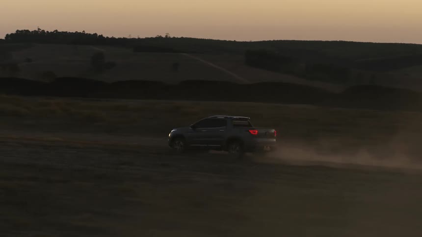 A pickup truck traveling on a dirt road near green fields leaves a trail of dust