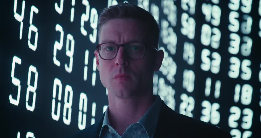 A man in glasses looks intensely into the camera as a screen behind him displays constantly changing numbers