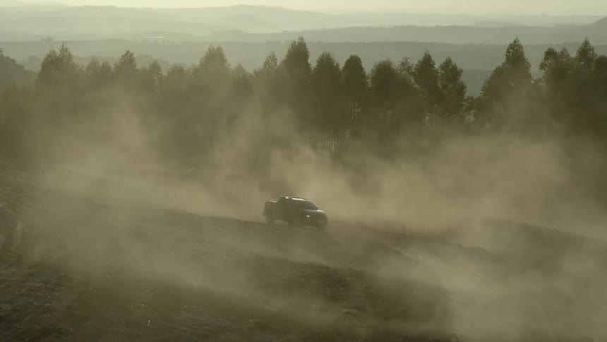 Aerial view of a pickup truck on a dirt slope surrounded by dust near a pine tree forest at evening