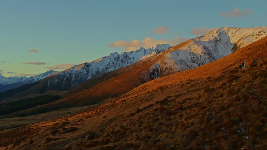 A drone flies over a wide open view of mountains with snow and hills with greenery