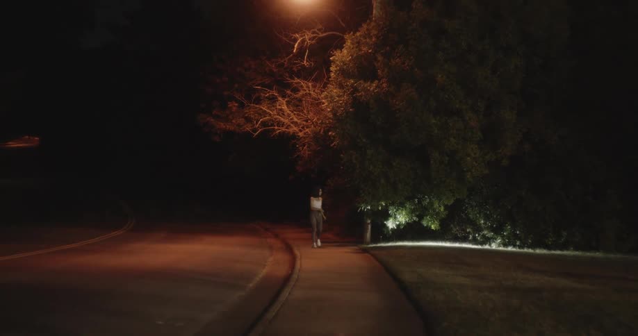 A woman is walking on a sidewalk at night with a flashlight looking for something near the trees