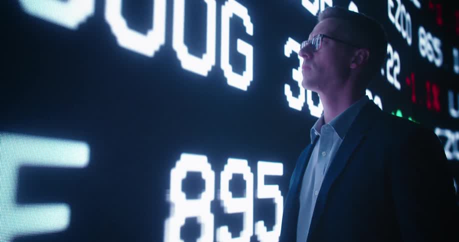 Young business man stands in front of a giant wall display looking at stock market values