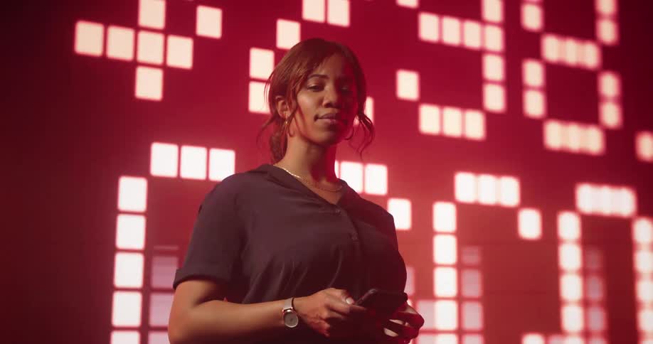 Young woman stands in front of a big digital display stock market values in a red hue
