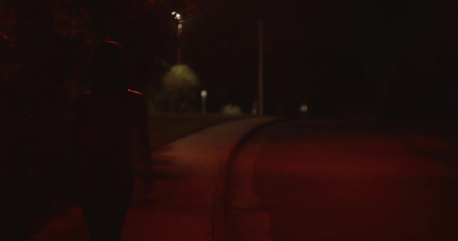 A woman is walking in the darkness on a sidewalk next to a street at night