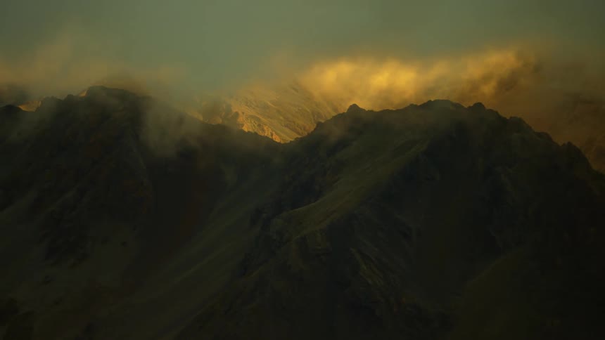 Clouds float over mountain peaks as the sun shines on a mountain ridge in the distance