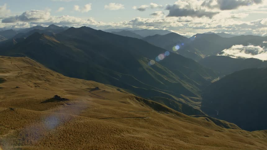 A trail winds down a brown slope towards a valley with mountain peaks and ridges on a sunny day