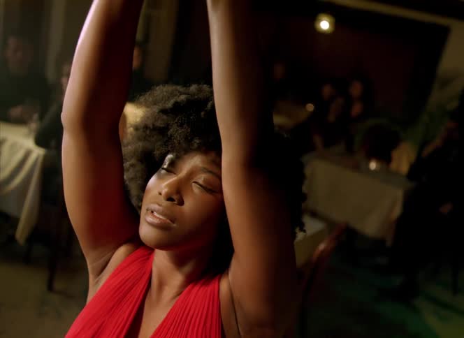 Light shines on the face of a woman standing in a restaurant moving her arms in the air and dancing as people sit at tables behind her