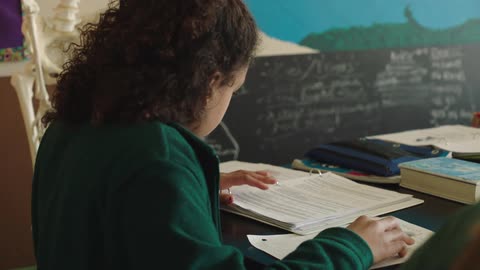 Girl with curly hair and green jacket sits at desk studying in room