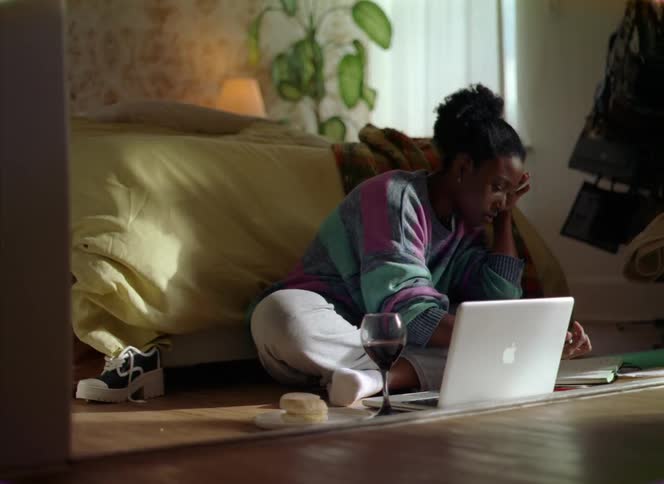 A woman is sitting on the floor with a laptop computer and a sandwich as she looks at a paper and types on the keyboard