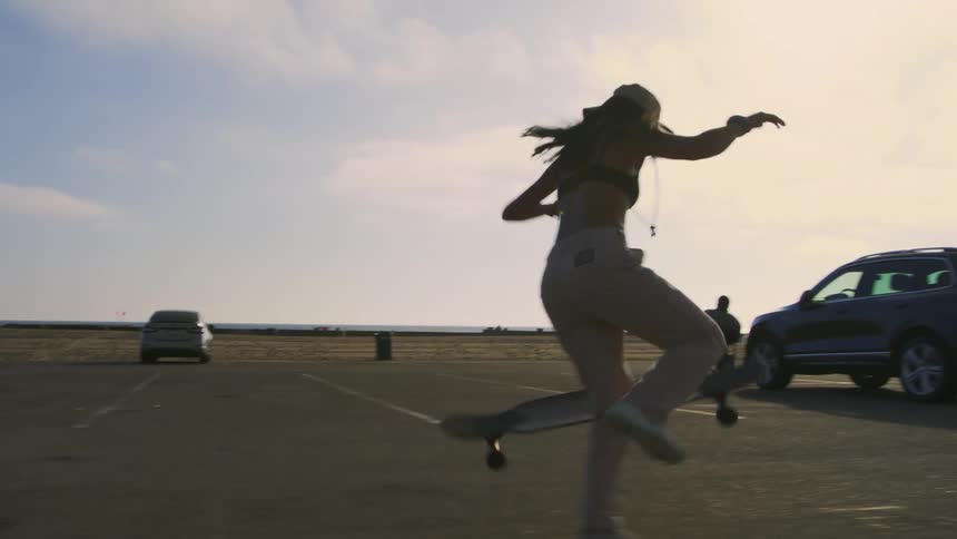 A female skateboarder does a trick while skating across the parking lot at a beach during the day