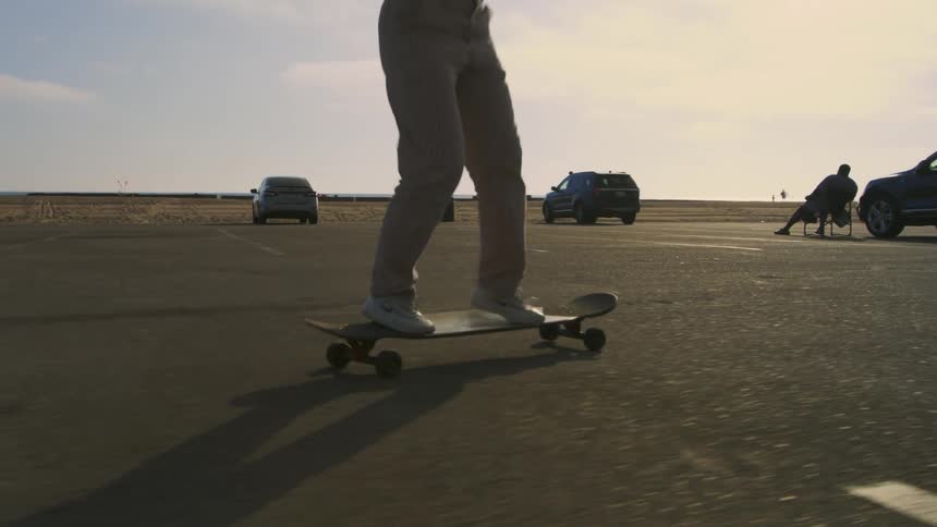 Woman with hat black top brown pants and white shoes skateboards in car park next to beach