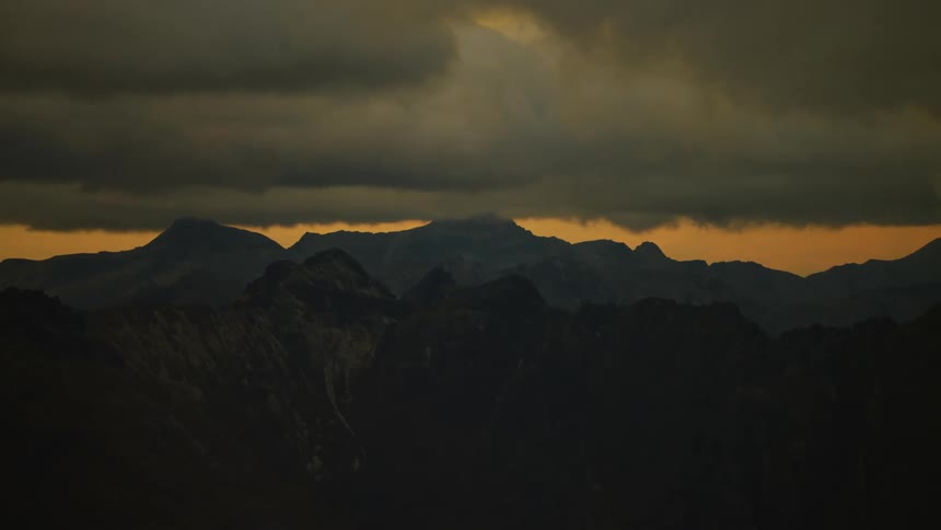 Dark clouds float over mountains with an orange sky in the distance