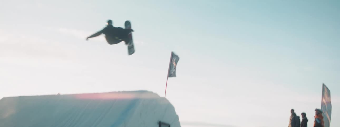 A man snowboards down the side of a mountain and flips over a jump