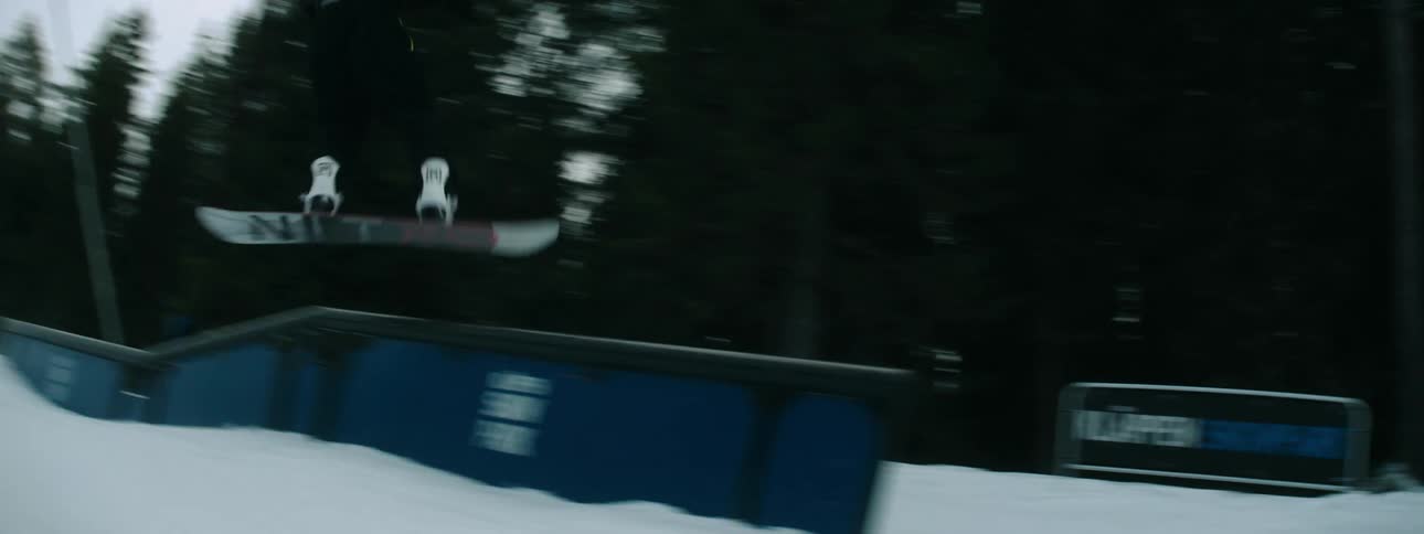 Snowboarder performs jump and spin before riding down snow covered slope toward buildings