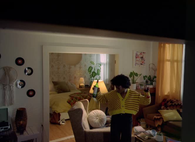 Woman with curly hair knit jacket holds green wine bottle and glass dancing in living room at home