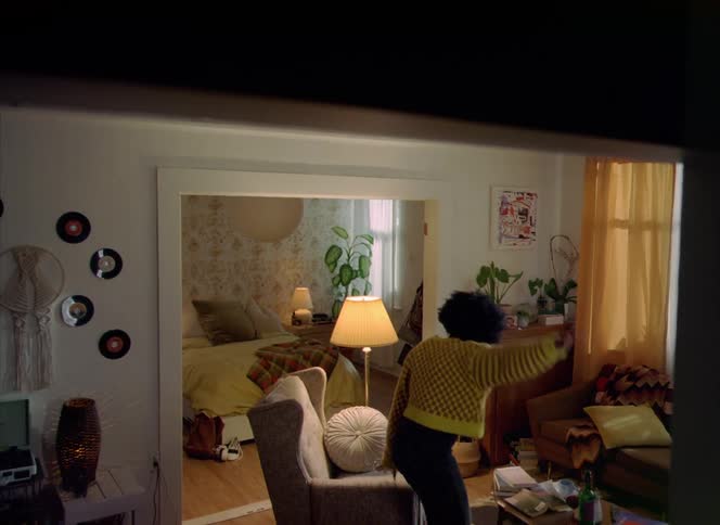Woman with curly hair knit jacket holds green wine bottle and glass dancing in living room at home