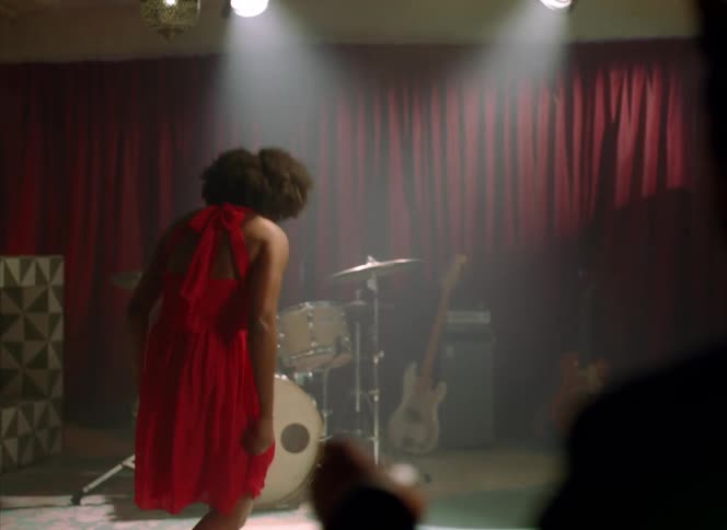 Man watches female dancer with curly hair and red dress on stage with drum kit and guitar in front of red curtains