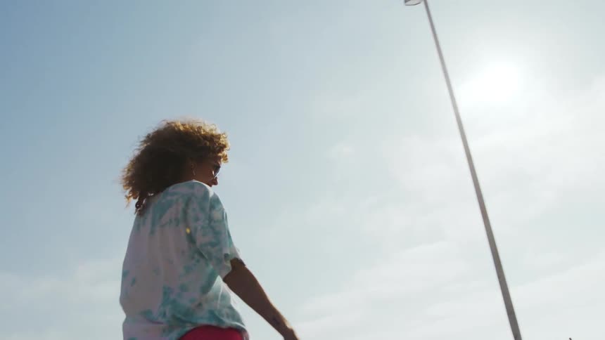 Woman with curly hair sunglasses shirt and pink shorts skateboards in car park next to beach