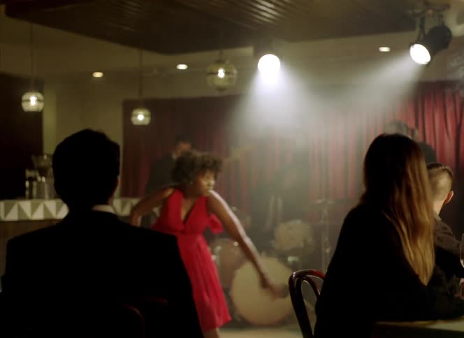 Barefoot woman wearing a red dress dances in front of a band on a restaurant