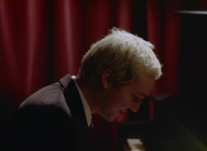 Blonde musician plays the piano energetically on stage in front of a red curtain