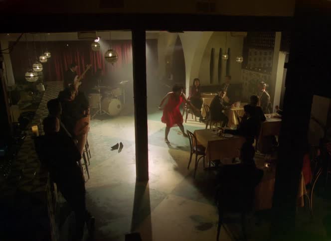 People watch female dancer with curly hair and red dress on stage with drum kit and guitar in front of red curtain in bar