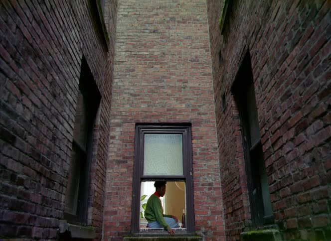 Woman with curly hair green sweater and jeans sits at windowsill in apartment building with brick wall