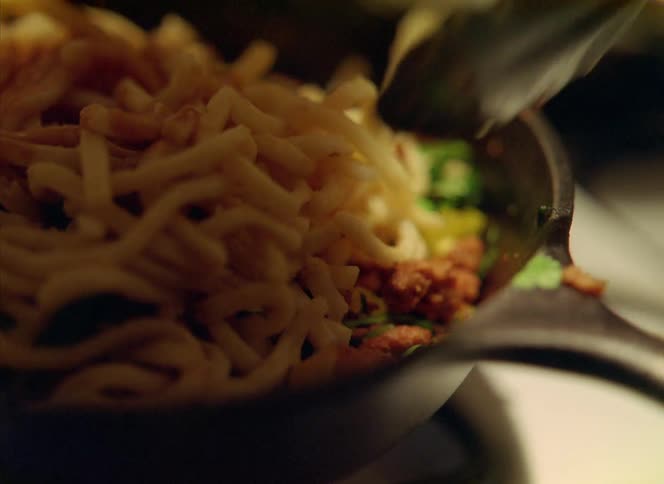 Someone is cooking and puts noodles on top of meat and vegetables in a frying pan and they are stirring it with tongs