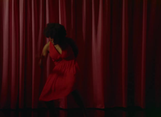 A woman wearing a red dress is dancing on a stage in front of a red curtain