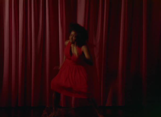 Woman with curly hair and red dress dances in front of red curtains on the stage
