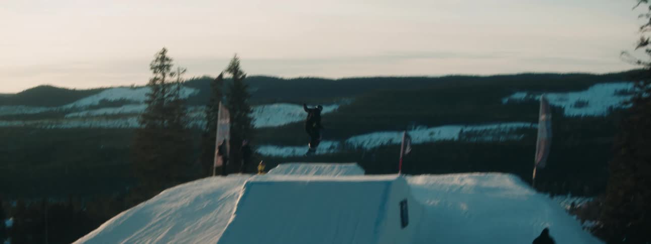 Man rides a snowboard down a slope to jump over a ramp in the mountains at sunset