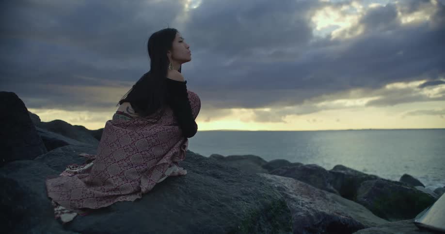 A woman kneels on rocks by the sea at sunset