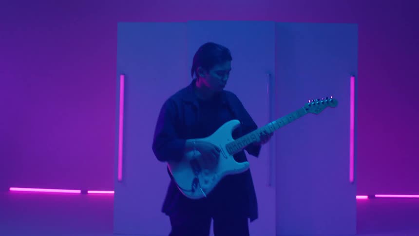Guitarist plays instrument against panel decorated with neon bars in area lit by colored lighting