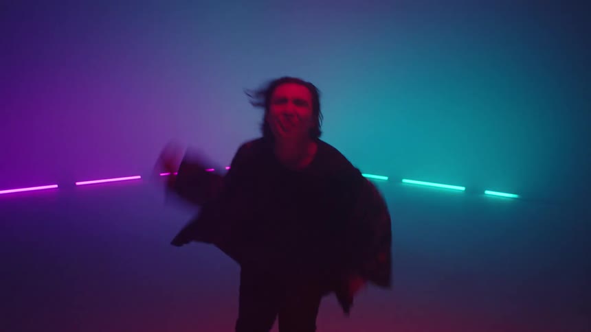 A man is singing and dancing as colored lights flash in the foggy air behind him
