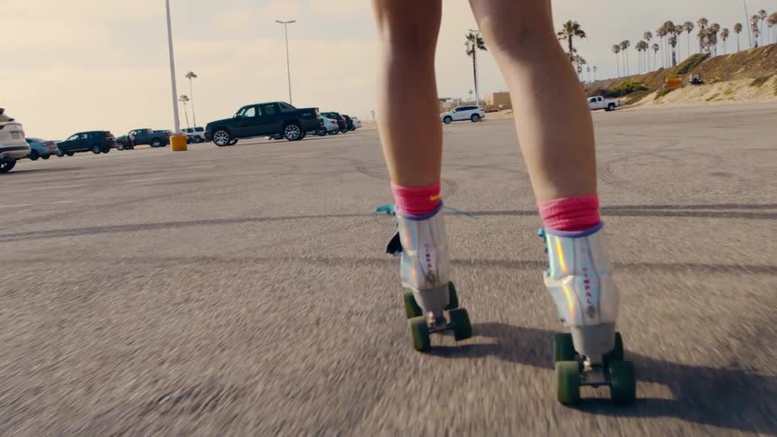 A girl is roller skating in a parking lot with cars parked and trees in the distance