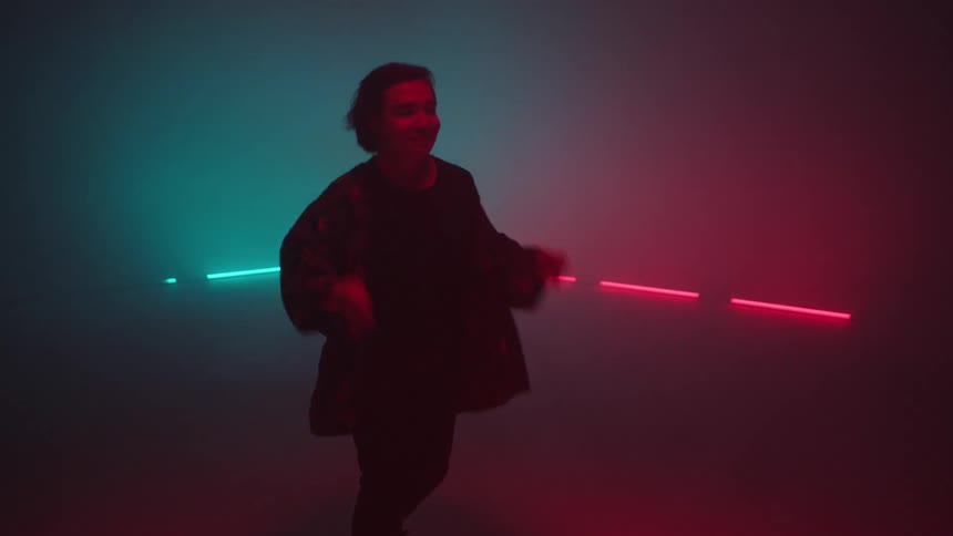 A man glances at the camera while dancing in front of colorful flashing lights in a foggy room