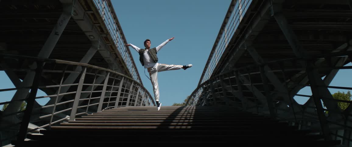 A man is standing on an outdoor staircase and as he lifts his arms and strikes a pose on one foot he floats into the air