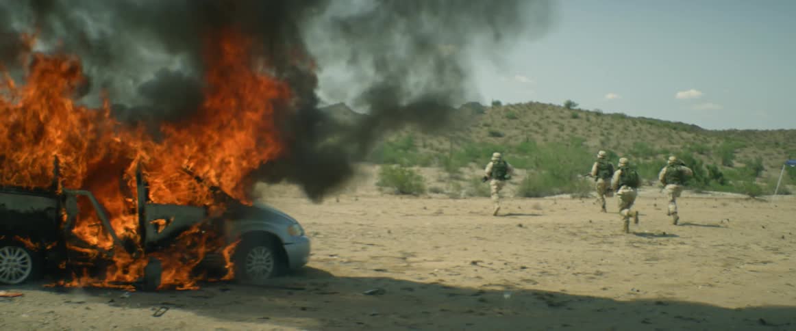 Four soldiers carrying weapons run past a burning car towards a ridge