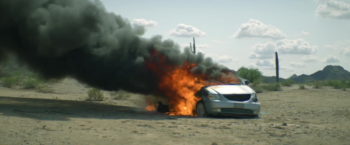 Thick black smoke rises from an automobile engulfed in flames in the desert