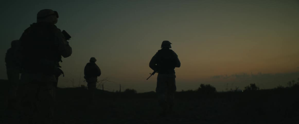Patrol of soldiers walk across field silhouetted against a red tinted sky