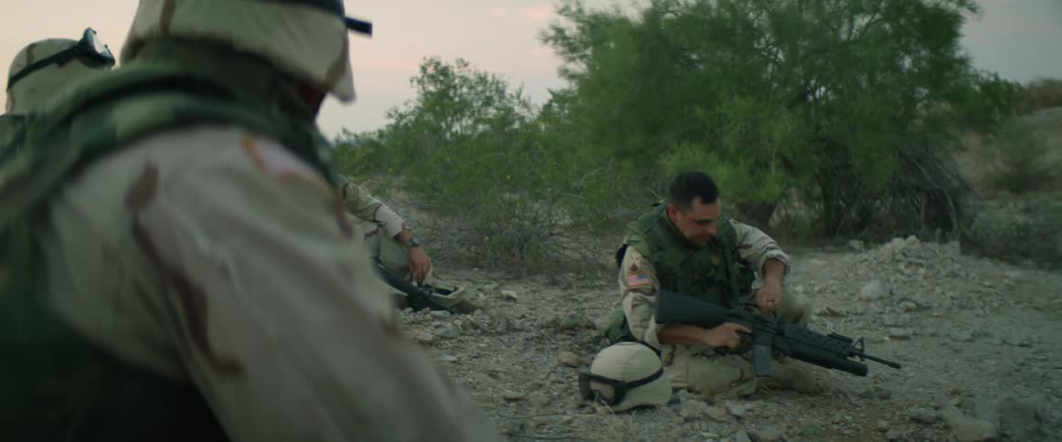 Soldiers carrying weapons sit on the ground talking to each other on at evening