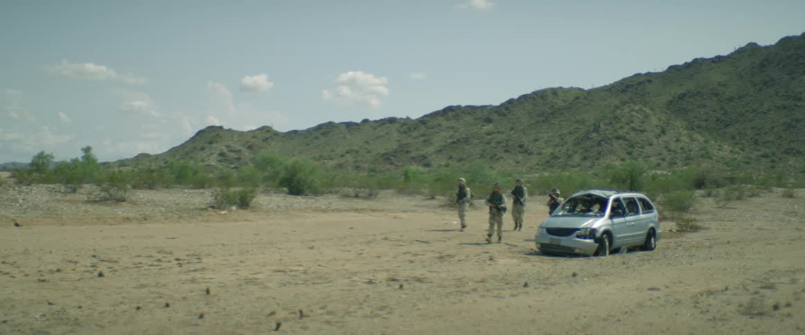 A small group of military soldiers are walking a near an abandoned automobile in the desert