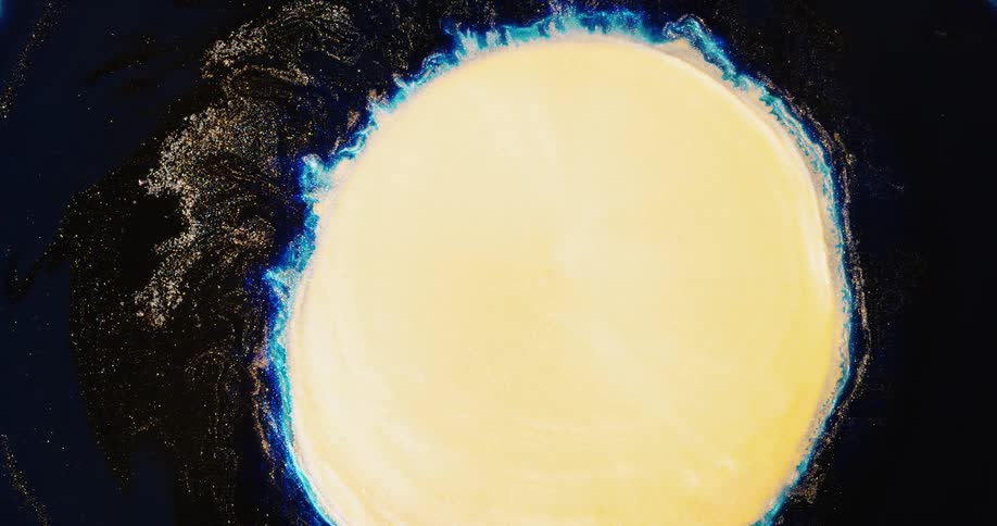 A blue burst of colored light explodes in a round shape as light yellow grows in from the center and the organic object shrinks as gold glitter sparkles around it
